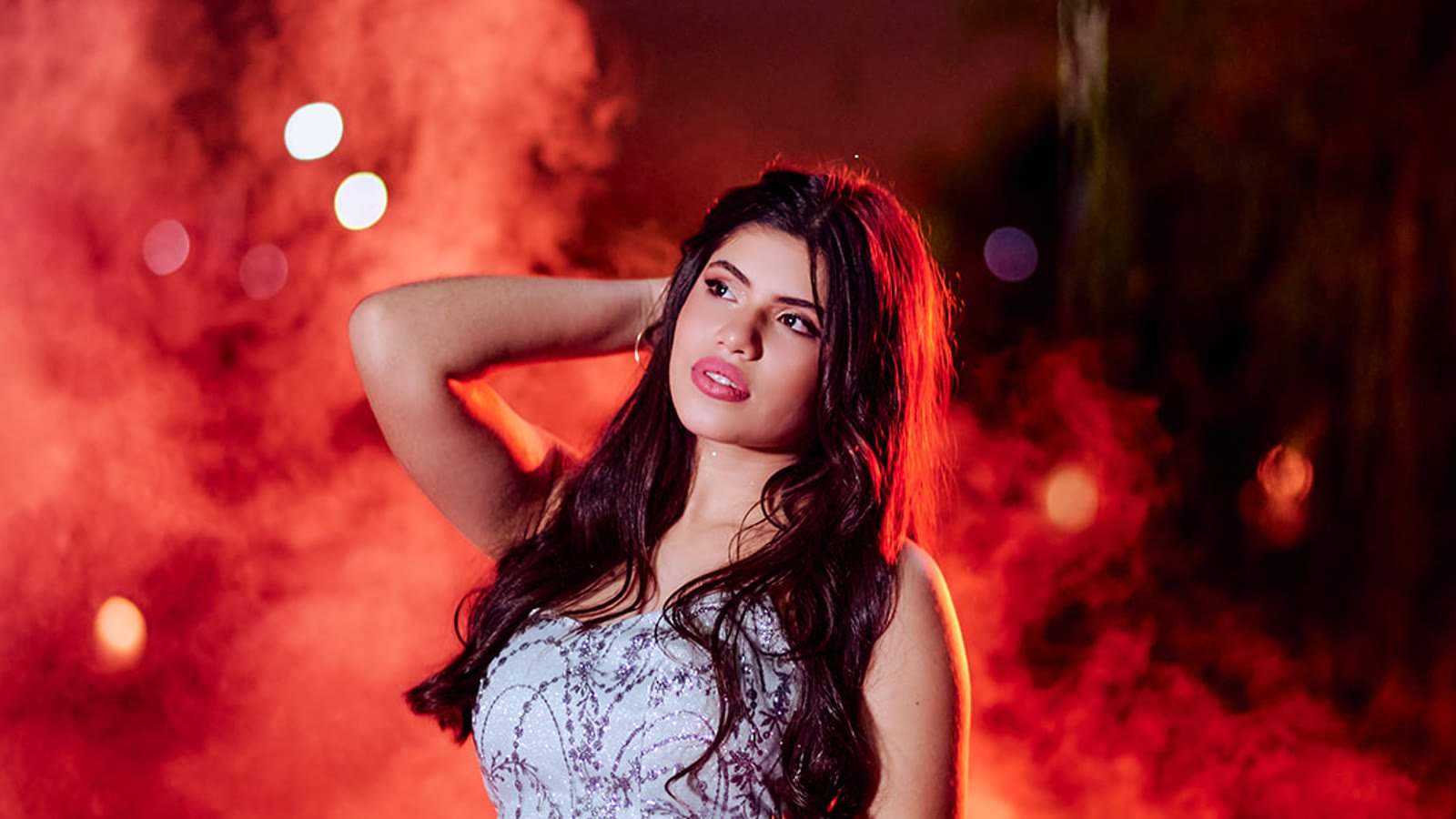 Quinceañera con conjunto plateado bordado, rodeada de humo rojo iluminado en una sesión nocturna
