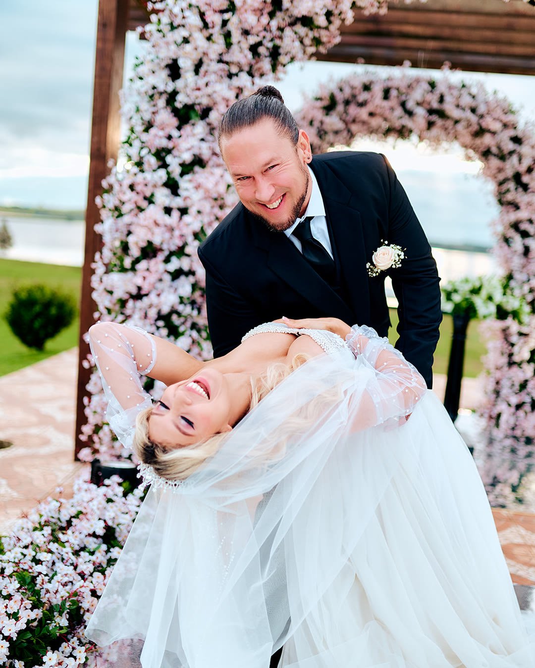 Novio inclinado sonriendo a la cámara mientras la novia se ríe a carcajadas, frente a un arco de flores rosadas