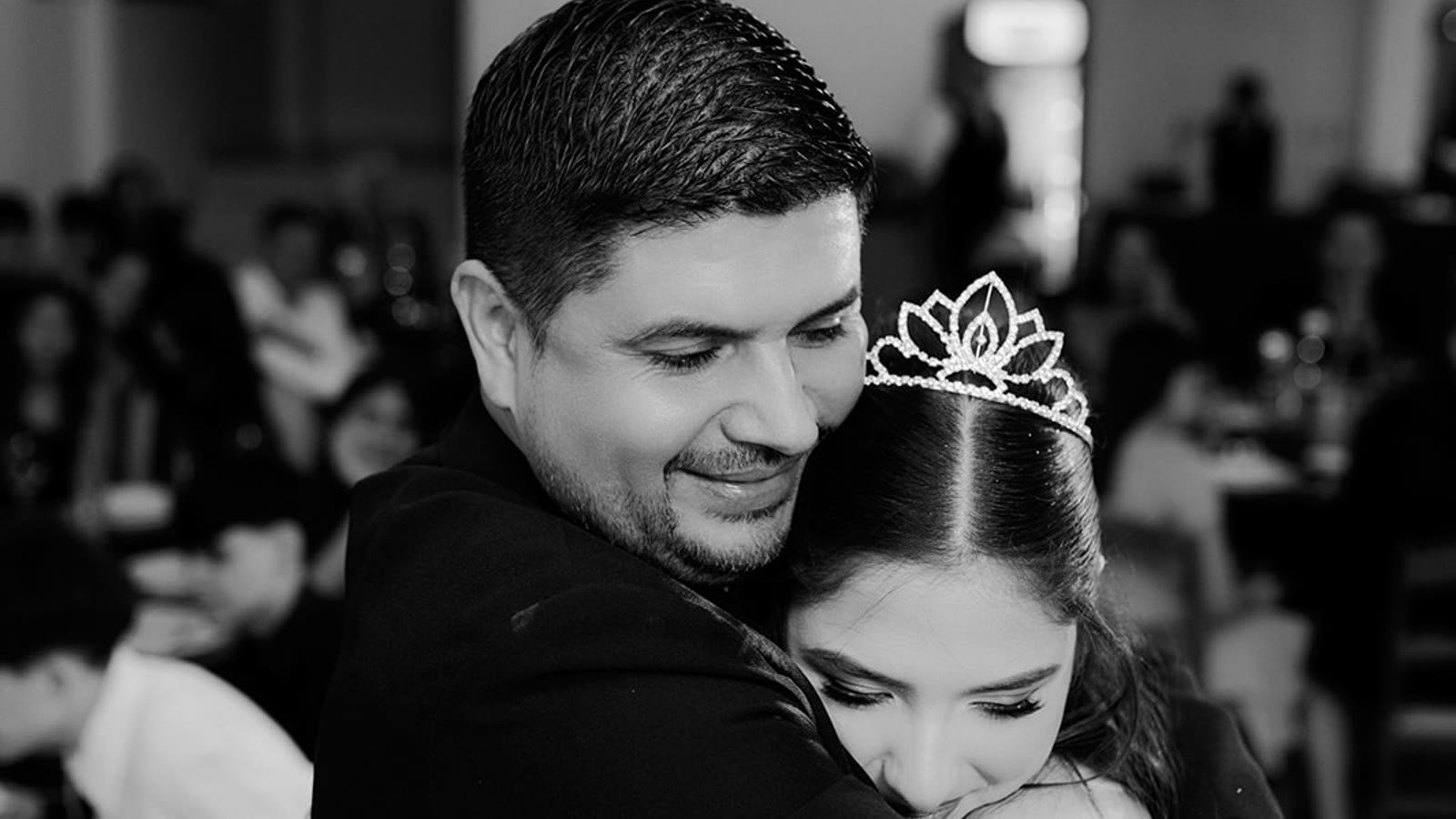 Un papá abraza a su hija quinceañera con tiara durante el vals, en blanco y negro, con los invitados desenfocados detrás