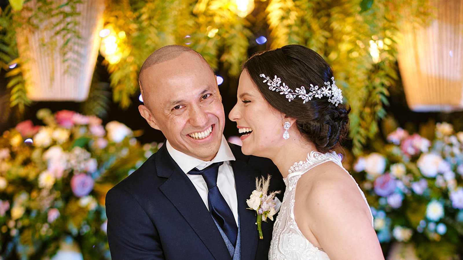 Novios riéndose muy cerca uno del otro, frente a frente, rodeados de flores en su boda