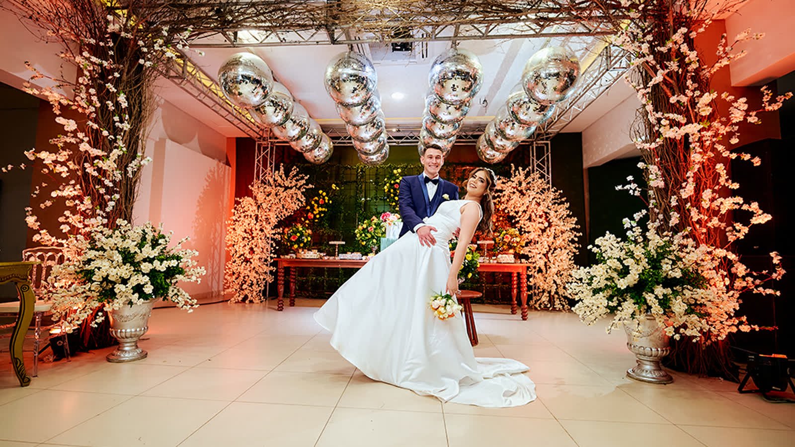 Novios abrazados en un salón decorado con flores blancas y esferas plateadas