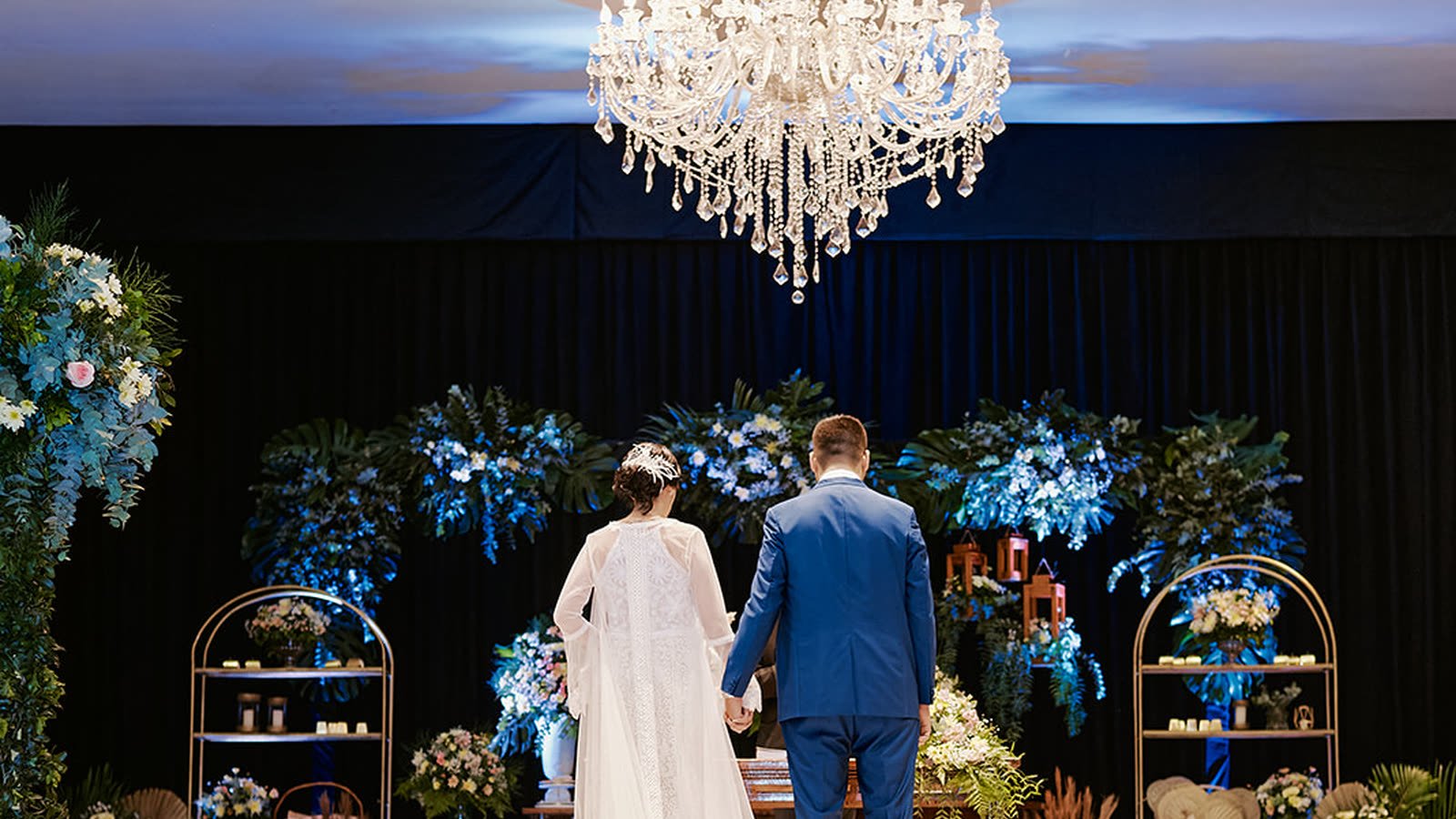 Novios de espaldas frente al altar decorado con flores, bajo una araña de cristal