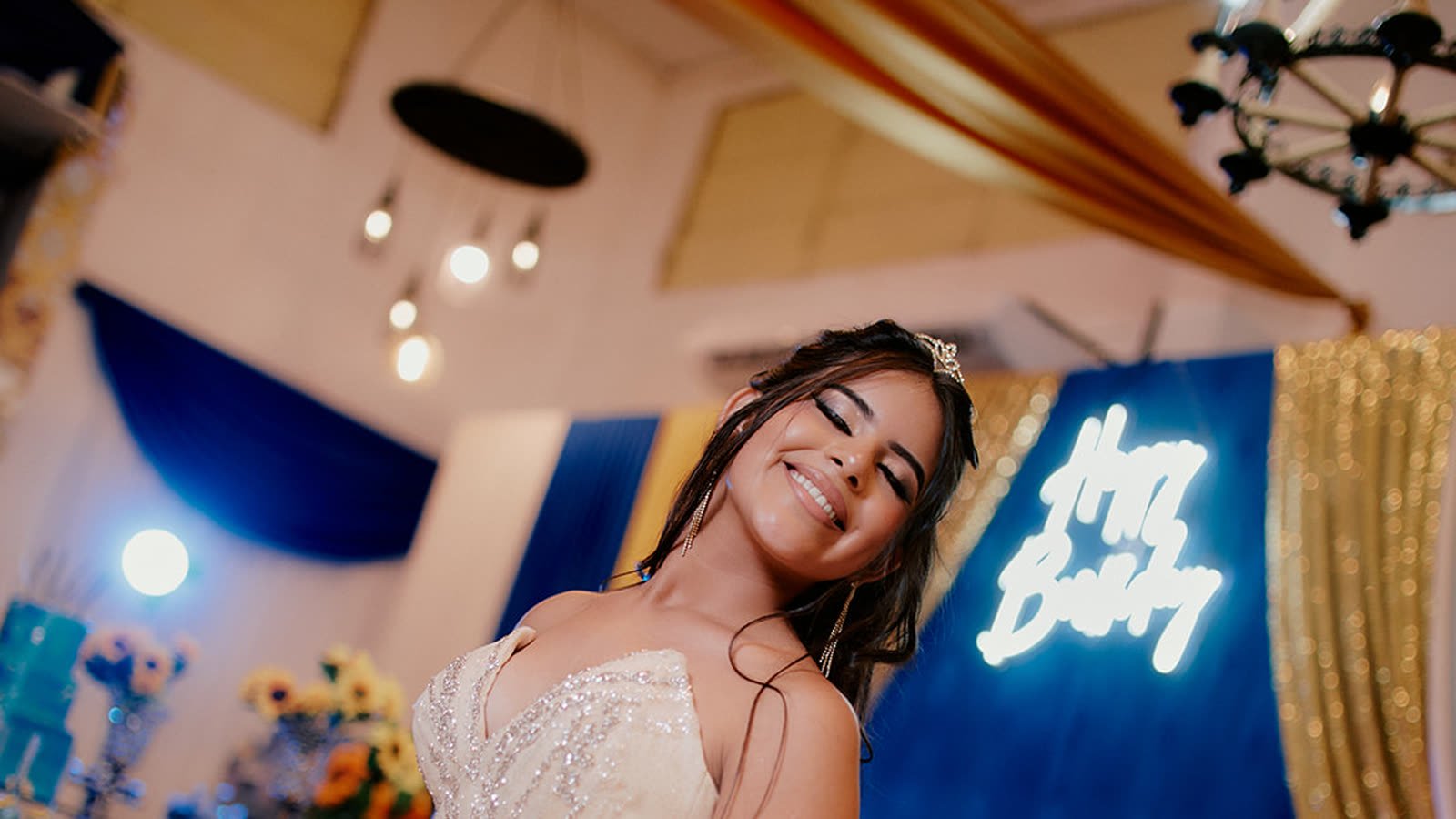 Quinceañera sonriendo con corona frente a un cartel de neón en su fiesta