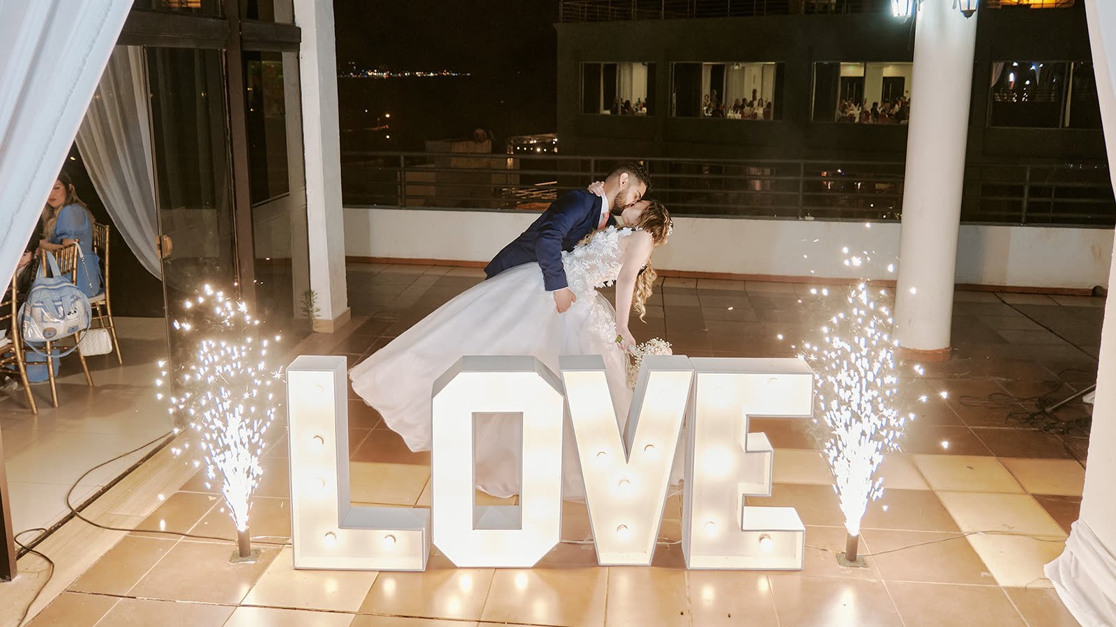 Novios besándose junto a letras luminosas LOVE con chispas frías en la fiesta