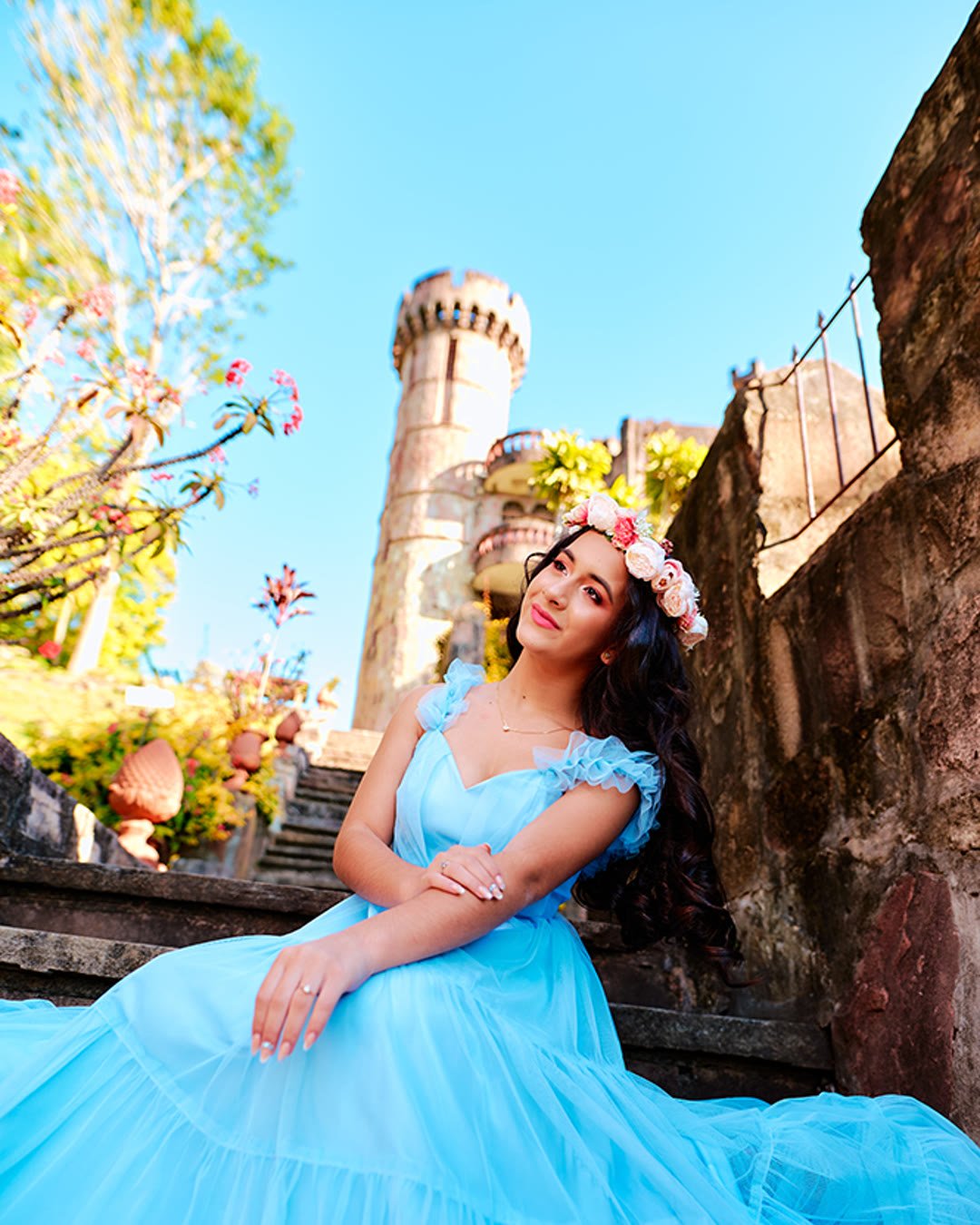 Quinceañera con vestido largo de tul celeste y corona de flores, sentada en una escalinata frente a una torre tipo castillo