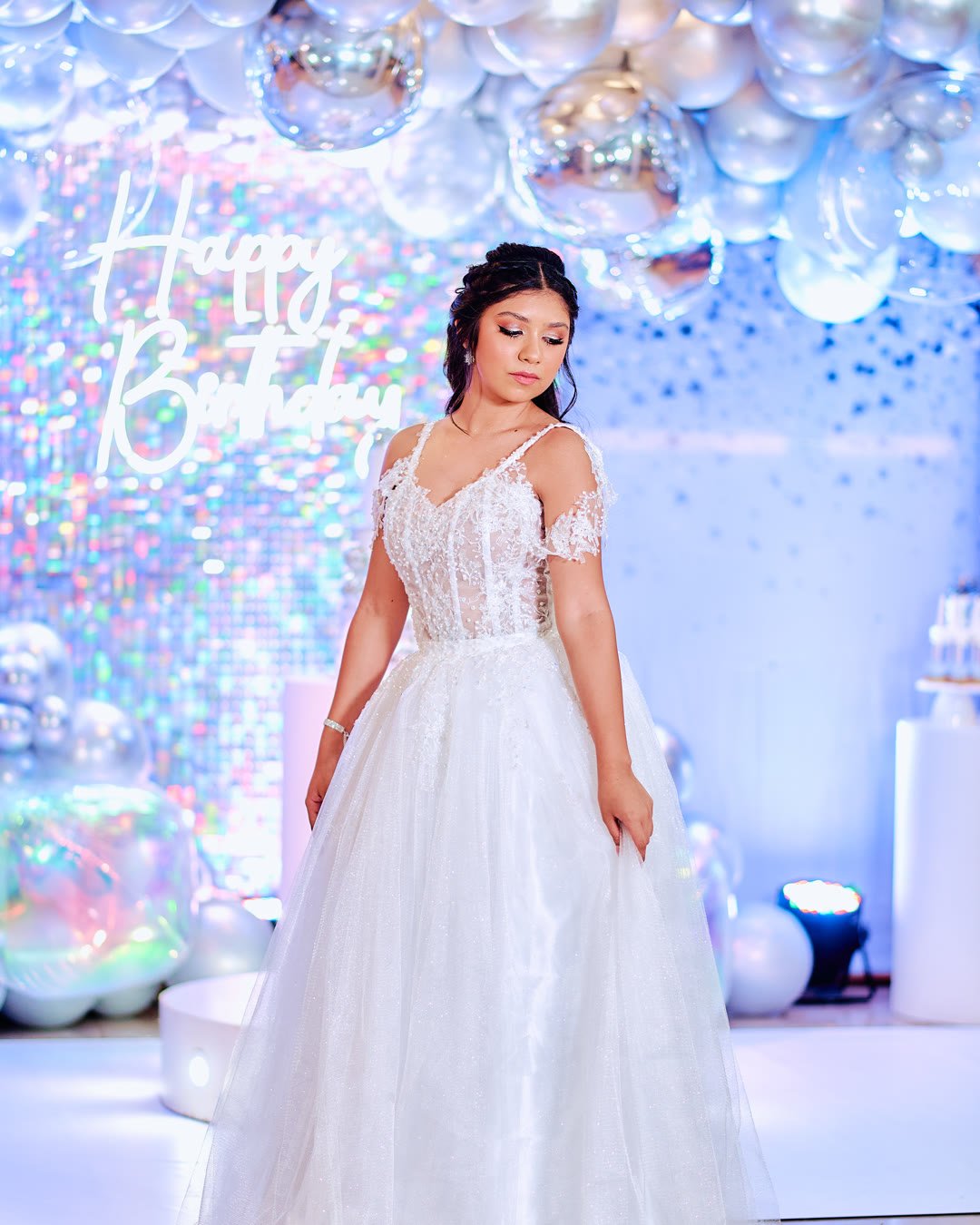 Quinceañera con vestido largo blanco de encaje en el salón de su fiesta, con globos plateados de fondo