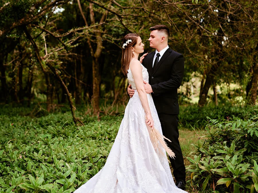 Novios abrazados en un bosque, ella con vestido de encaje y ramo de pampa