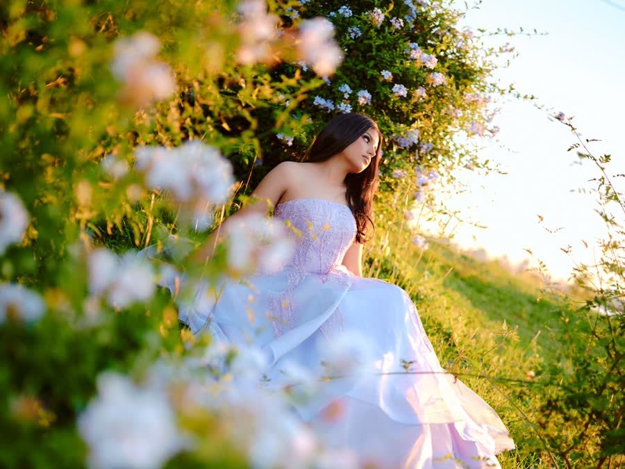 Quinceañera con vestido lila sentada entre flores al atardecer, en su sesión pre-xv