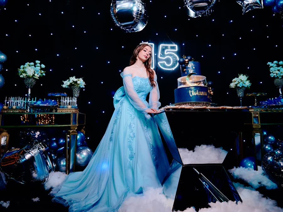 Quinceañera con vestido celeste junto a su torta y el número 15 iluminado en su fiesta