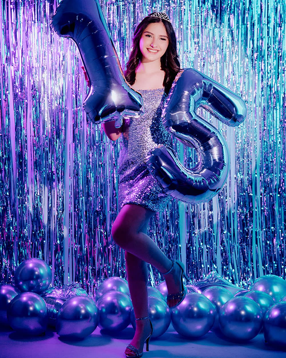 Quinceañera con globos plateados del número 15 frente a una cortina brillante